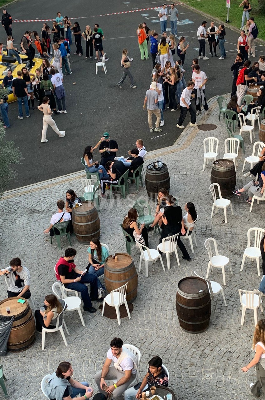 Groupe de personnes assises et debout autour de tonneaux et de chaises sur une place pavée, ambiance conviviale en extérieur.