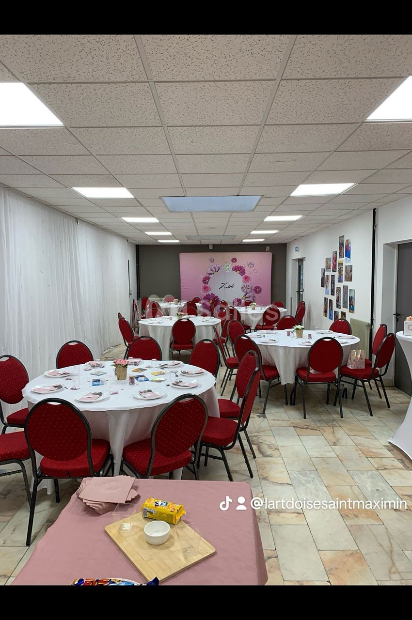 Salle décorée avec des tables rondes, nappes blanches et chaises rouges, prête pour un événement festif.