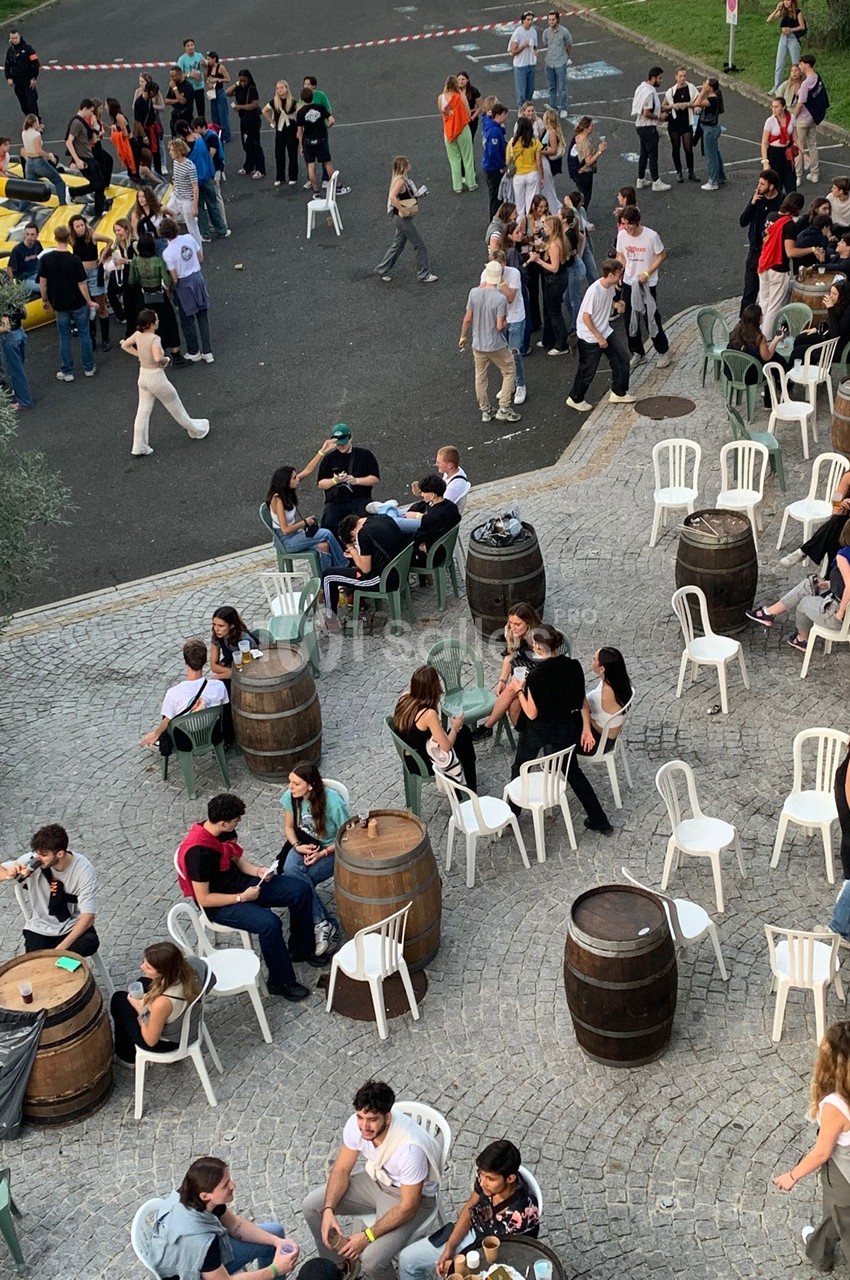 Groupe de personnes assises et debout lors d'un événement en plein air, entourées de tables et tonneaux sur une place pavée.
