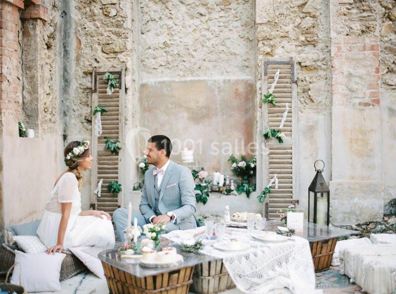 Un couple assis dans une cour rustique décorée pour un mariage, entouré de tables basses et de détails floraux.