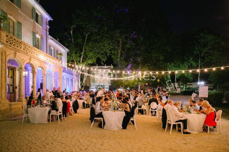 Dîner en plein air de nuit avec des invités assis à des tables rondes, éclairé par des guirlandes lumineuses.