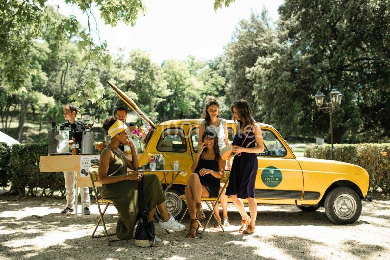 Un groupe de personnes discute et se détend près d'une voiture jaune transformée en stand de boissons dans un parc.