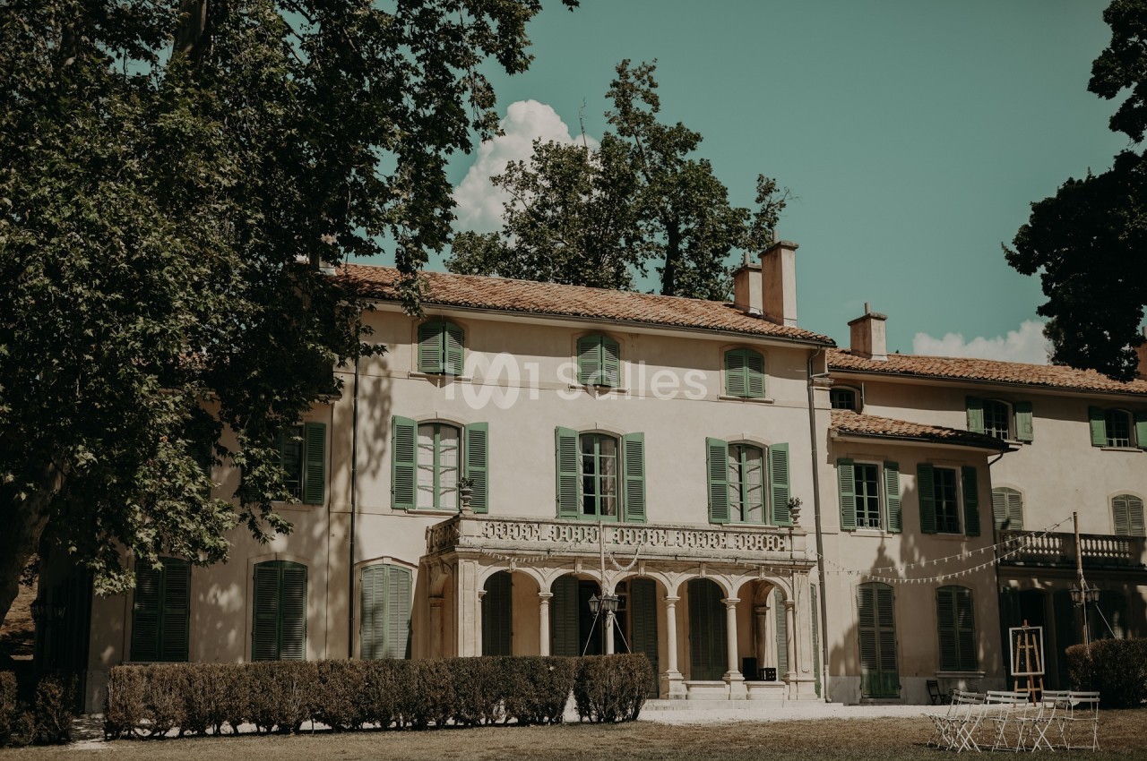 Façade d'une grande maison ancienne avec volets verts, entourée d'arbres et d'une pelouse.