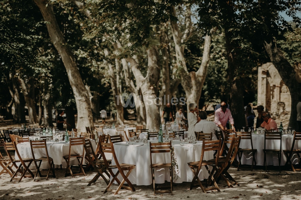 Tables rondes dressées avec nappes blanches et chaises en bois disposées en extérieur sous des arbres.
