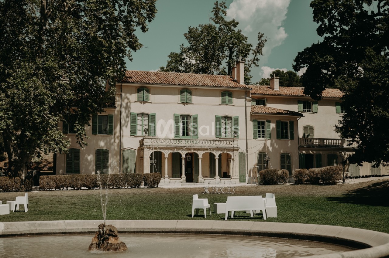 Façade d'une grande maison provençale entourée d'arbres, avec une fontaine et des bancs blancs au premier plan.