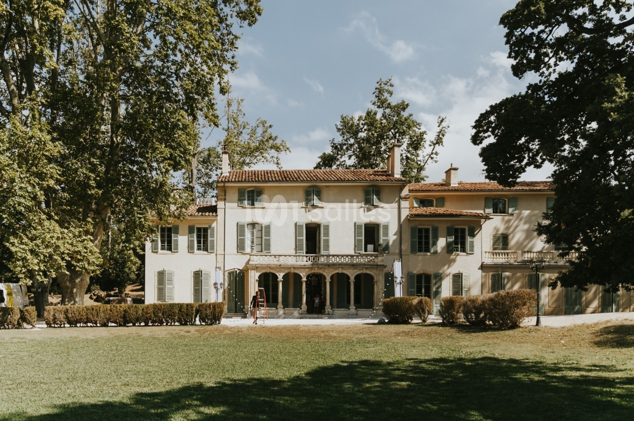 Grande maison provençale entourée d'arbres, avec une façade claire et un toit en tuiles rouges, vue depuis un jardin.