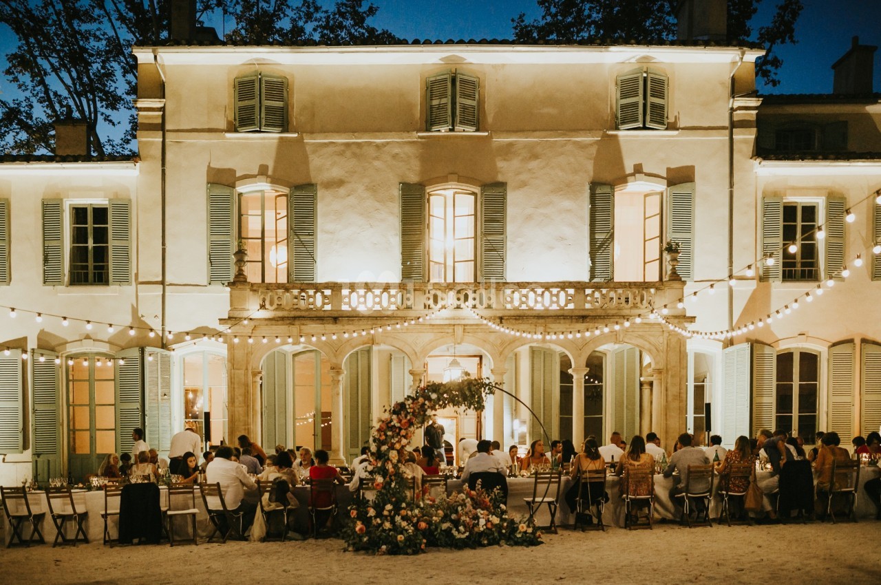Dîner en plein air devant une grande bâtisse illuminée, avec des invités assis à une longue table décorée de fleurs.