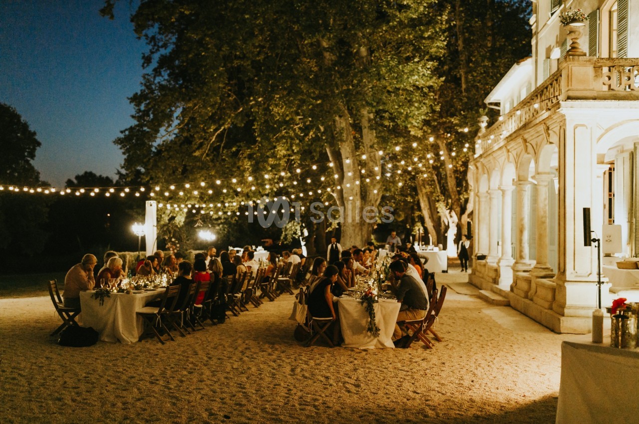 Dîner en plein air sous des guirlandes lumineuses, avec des invités assis à des tables près d'un bâtiment élégant.