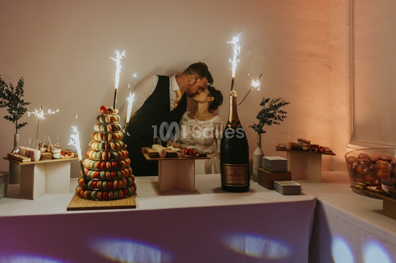 Un couple de mariés s'embrasse devant une table décorée avec des desserts, une pyramide de macarons et des étincelles.