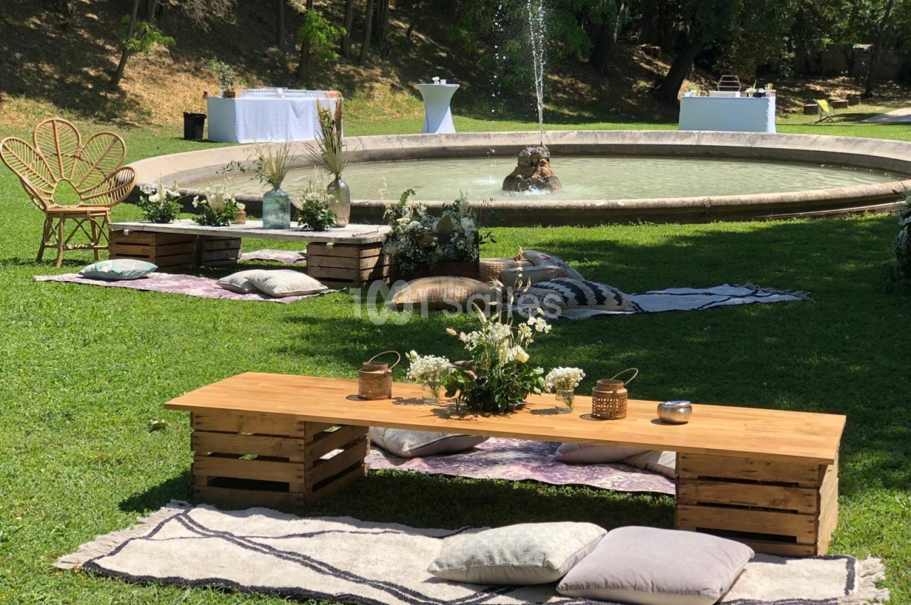 Décoration champêtre avec banc en bois, coussins et fontaine en arrière-plan dans un jardin verdoyant.