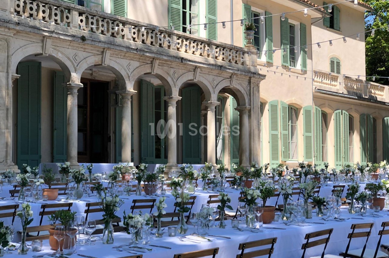 Tables dressées pour un repas en extérieur devant une grande bâtisse aux volets verts et à l'architecture classique.
