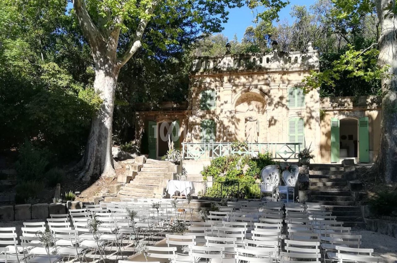 Chaises blanches alignées en extérieur devant une petite bâtisse ancienne entourée d'arbres et de verdure.