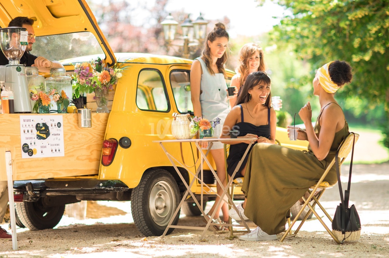 Deux femmes discutent à une table en extérieur près d'un food truck jaune, entourées de fleurs et de verdure.