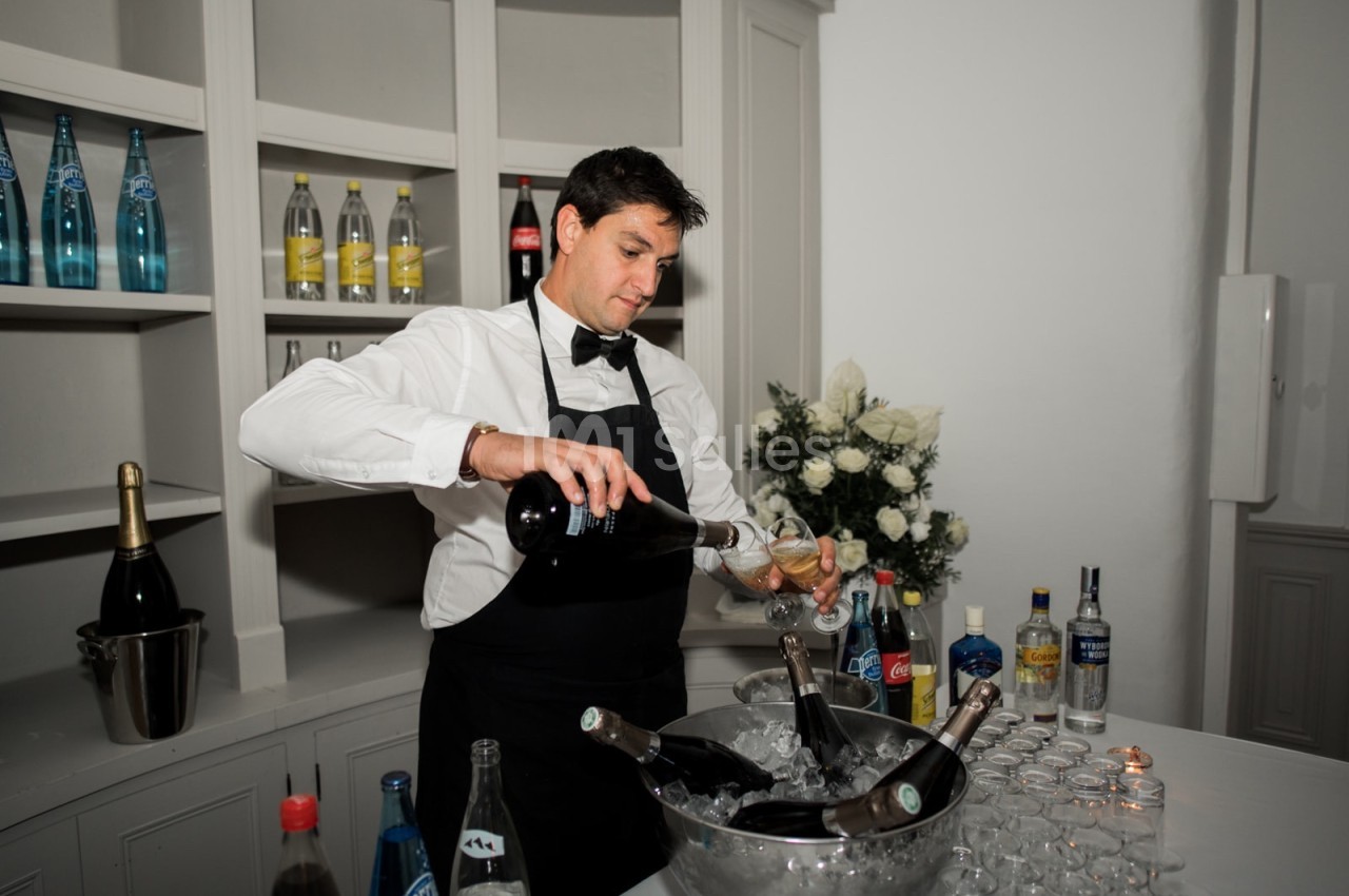 Un serveur en chemise blanche et tablier noir verse du champagne dans un verre devant un bar avec des bouteilles.