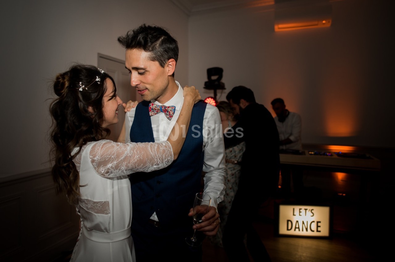 Un couple danse tendrement dans une salle faiblement éclairée, avec une pancarte lumineuse ’Let's Dance’ en arrière-plan.