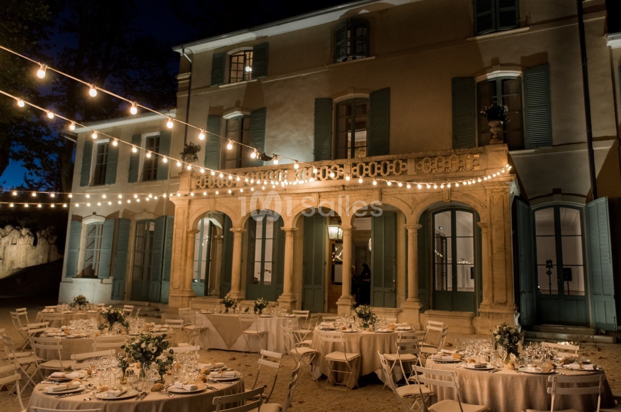 Façade d'un bâtiment éclairé la nuit, avec tables dressées et guirlandes lumineuses dans une cour extérieure.