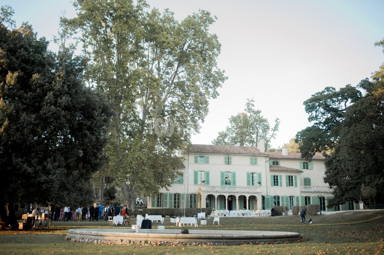 Grande maison provençale entourée d'arbres, avec une fontaine au premier plan et des invités réunis dans le jardin.