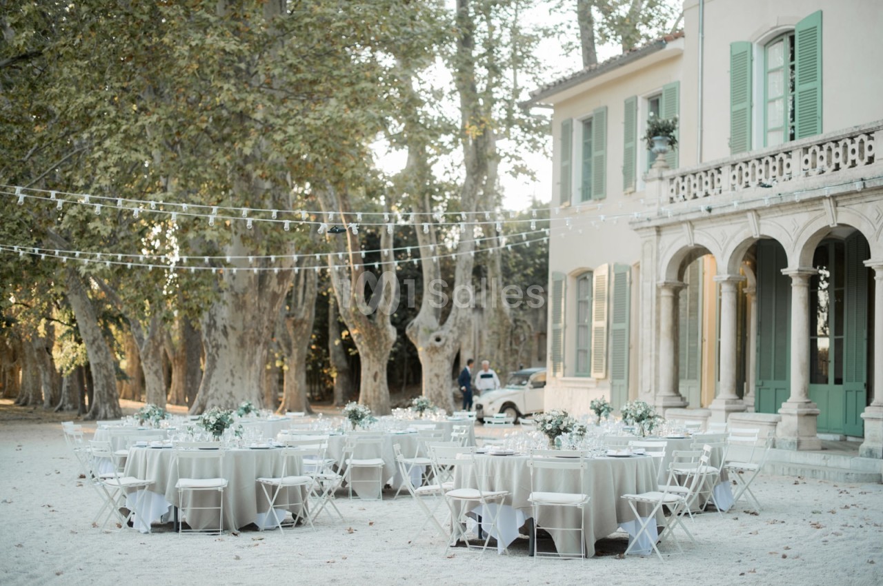Tables dressées pour un événement en plein air devant une grande maison avec des arbres et des guirlandes lumineuses.