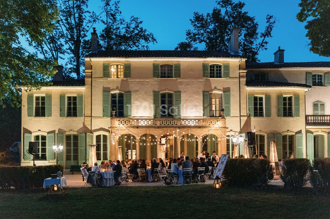Façade d'un manoir éclairé en soirée, avec des invités dînant en extérieur sous des guirlandes lumineuses.