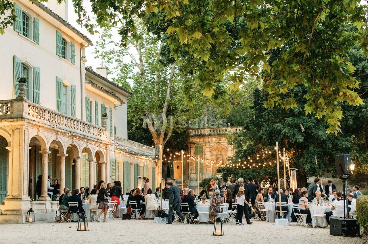 Dîner en plein air avec des invités assis à des tables rondes, devant une grande maison entourée d'arbres.