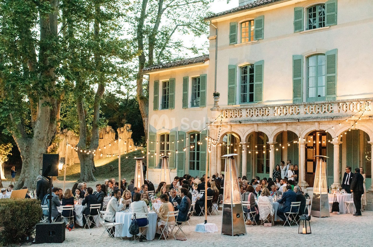 Dîner en plein air avec des invités assis à des tables, devant une grande maison avec des volets verts et des guirlandes…