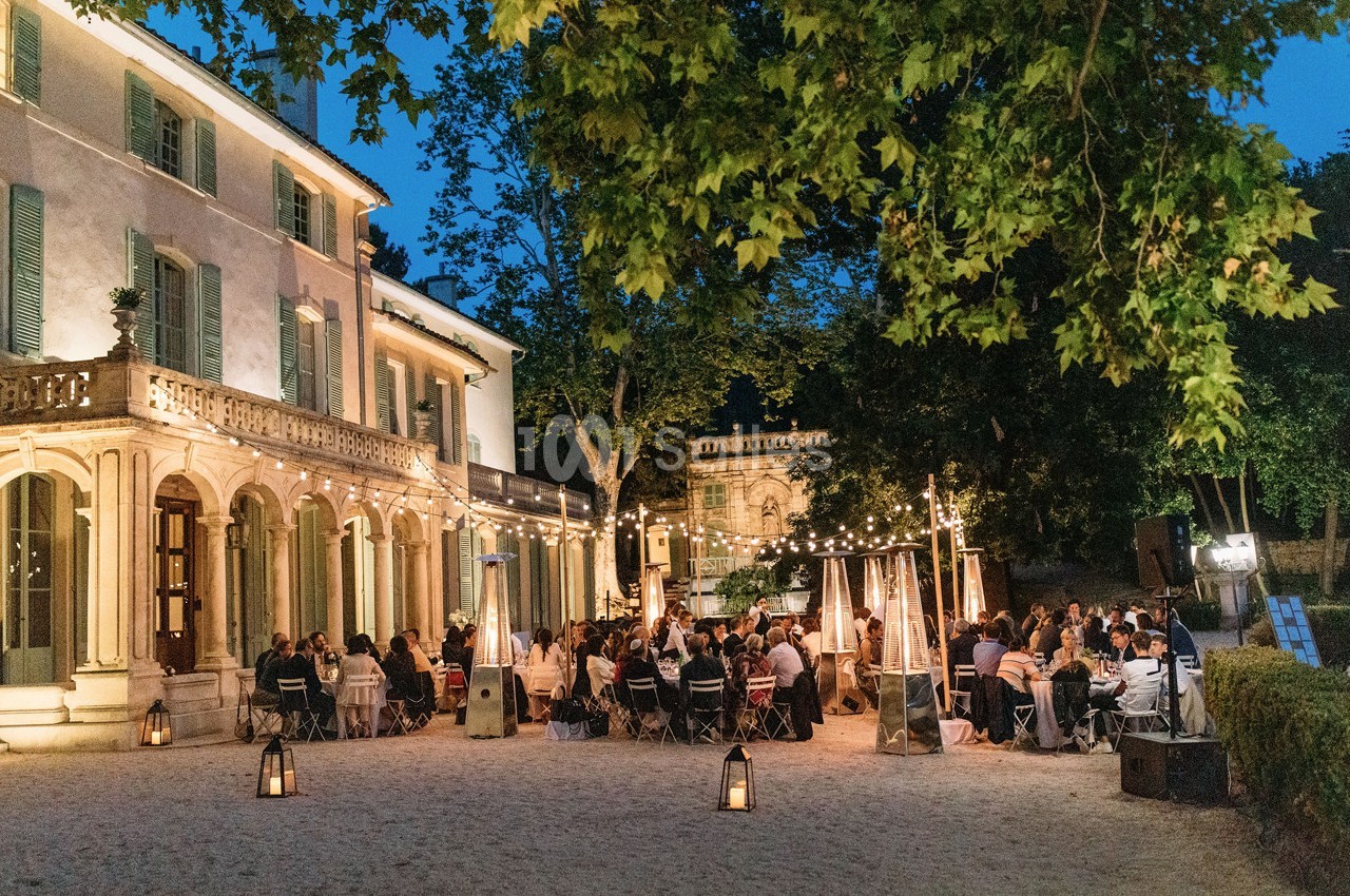 Dîner en plein air sous des guirlandes lumineuses devant un bâtiment ancien entouré d'arbres.