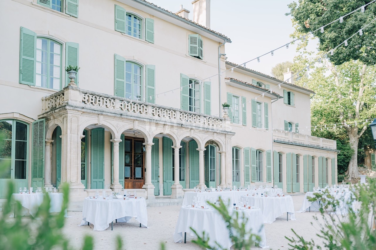 Façade d'une grande maison provençale avec volets verts, tables dressées en extérieur et guirlandes lumineuses.
