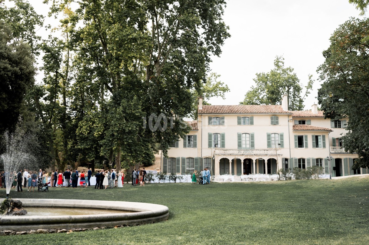 Manoir entouré d'un grand jardin verdoyant avec une fontaine au premier plan et des invités rassemblés à l'extérieur.