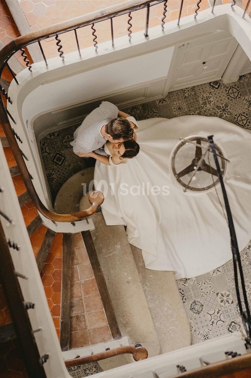 Vue plongeante d'un couple dans un escalier ancien, la femme porte une robe blanche ample.
