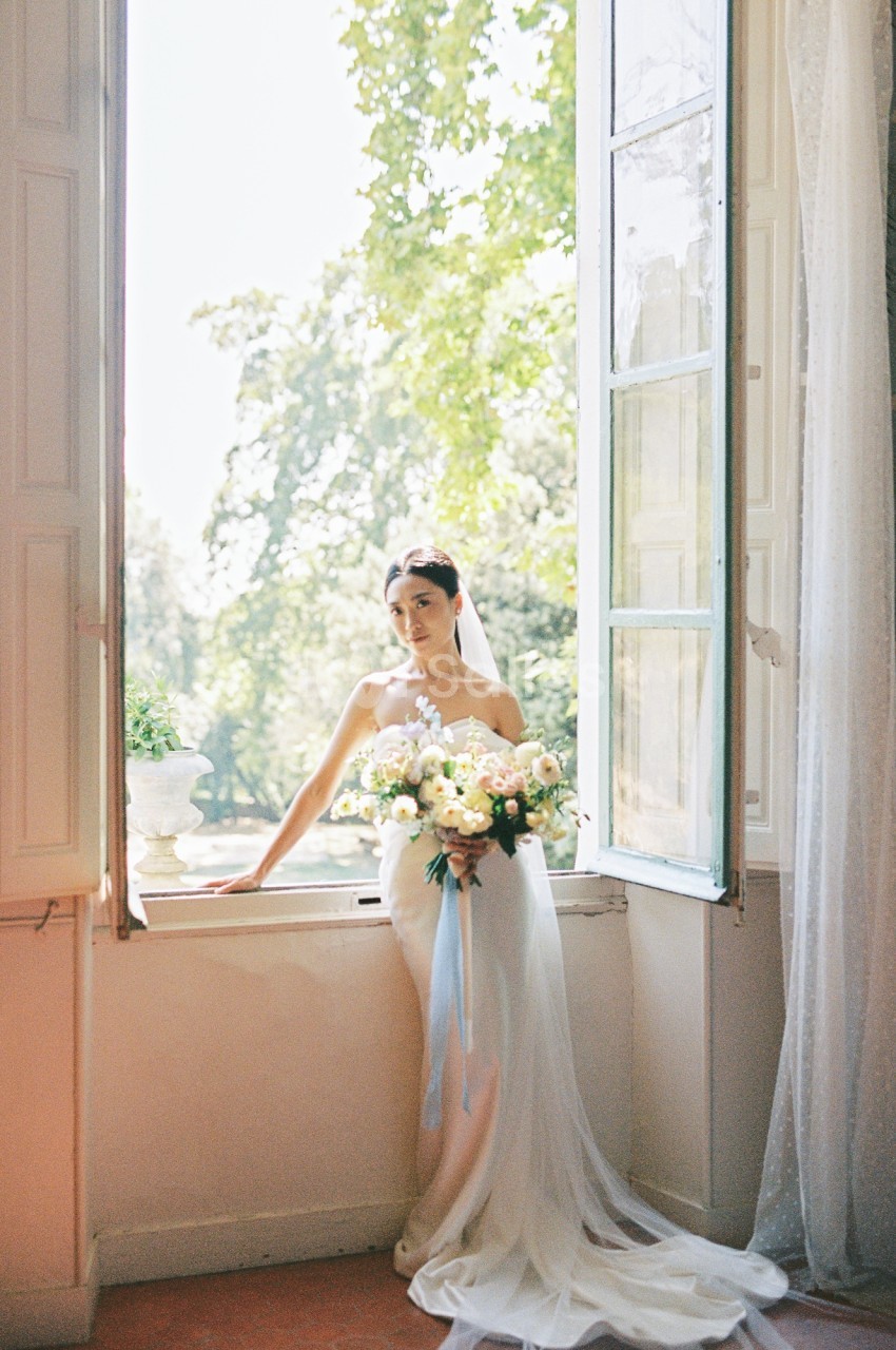 Une mariée en robe blanche tient un bouquet de fleurs devant une fenêtre ouverte avec vue sur des arbres verdoyants.