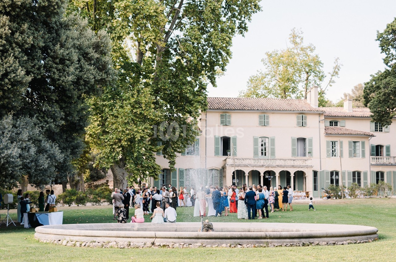 Groupe de personnes rassemblées près d'une fontaine dans un jardin, devant une grande maison ancienne entourée d'arbres.