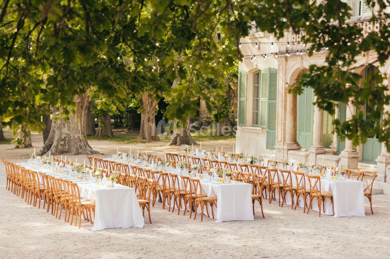 Tables longues décorées pour un repas en plein air, disposées devant un bâtiment ancien entouré d'arbres.