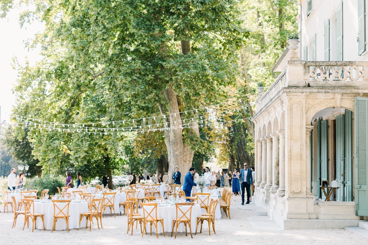 Tables et chaises en bois disposées en extérieur sous des guirlandes lumineuses près d'un bâtiment ancien entouré d'arbres.