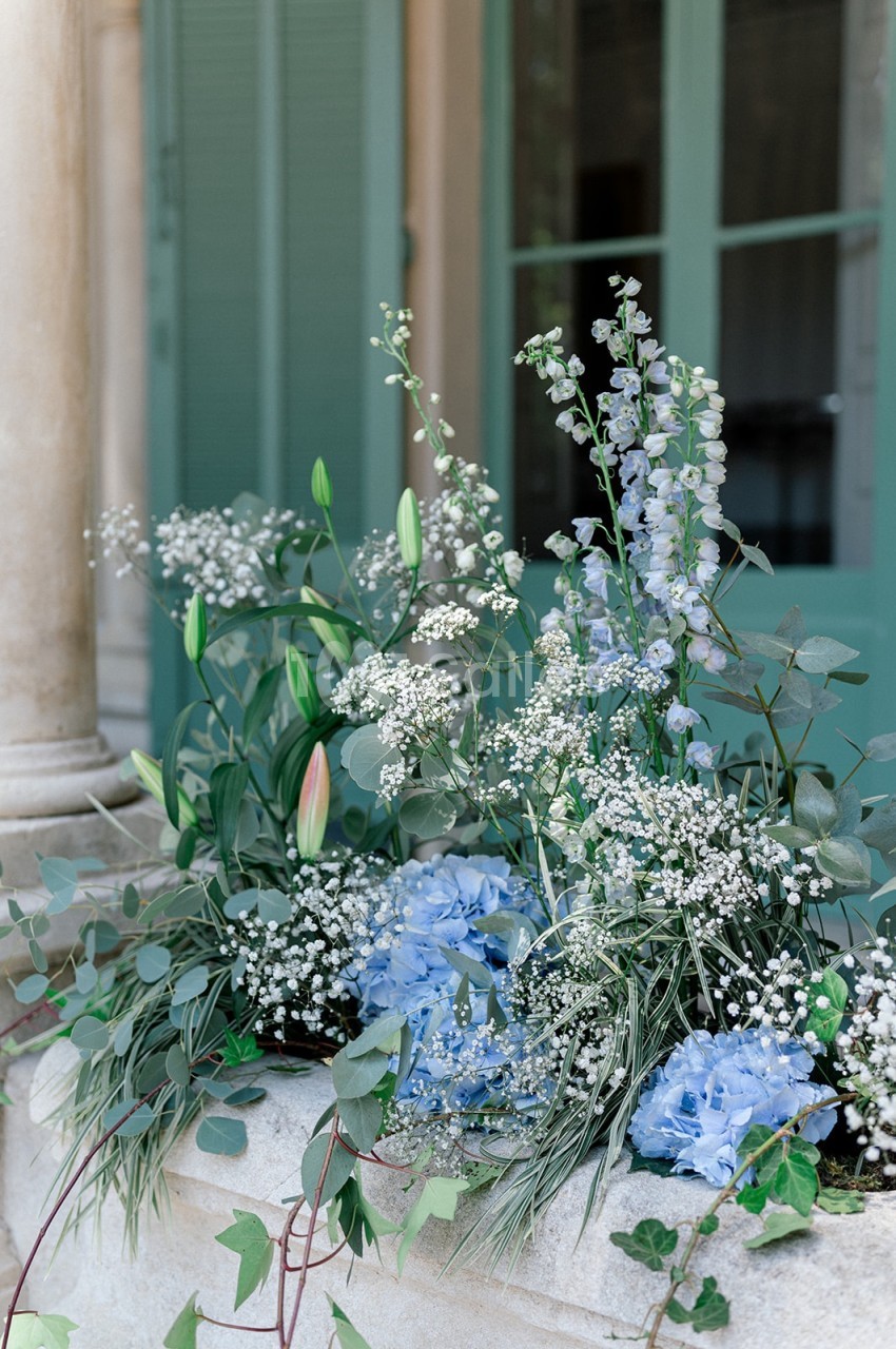 Composition florale avec hortensias bleus, gypsophiles, lys et feuillage, disposée devant une fenêtre à volets verts.