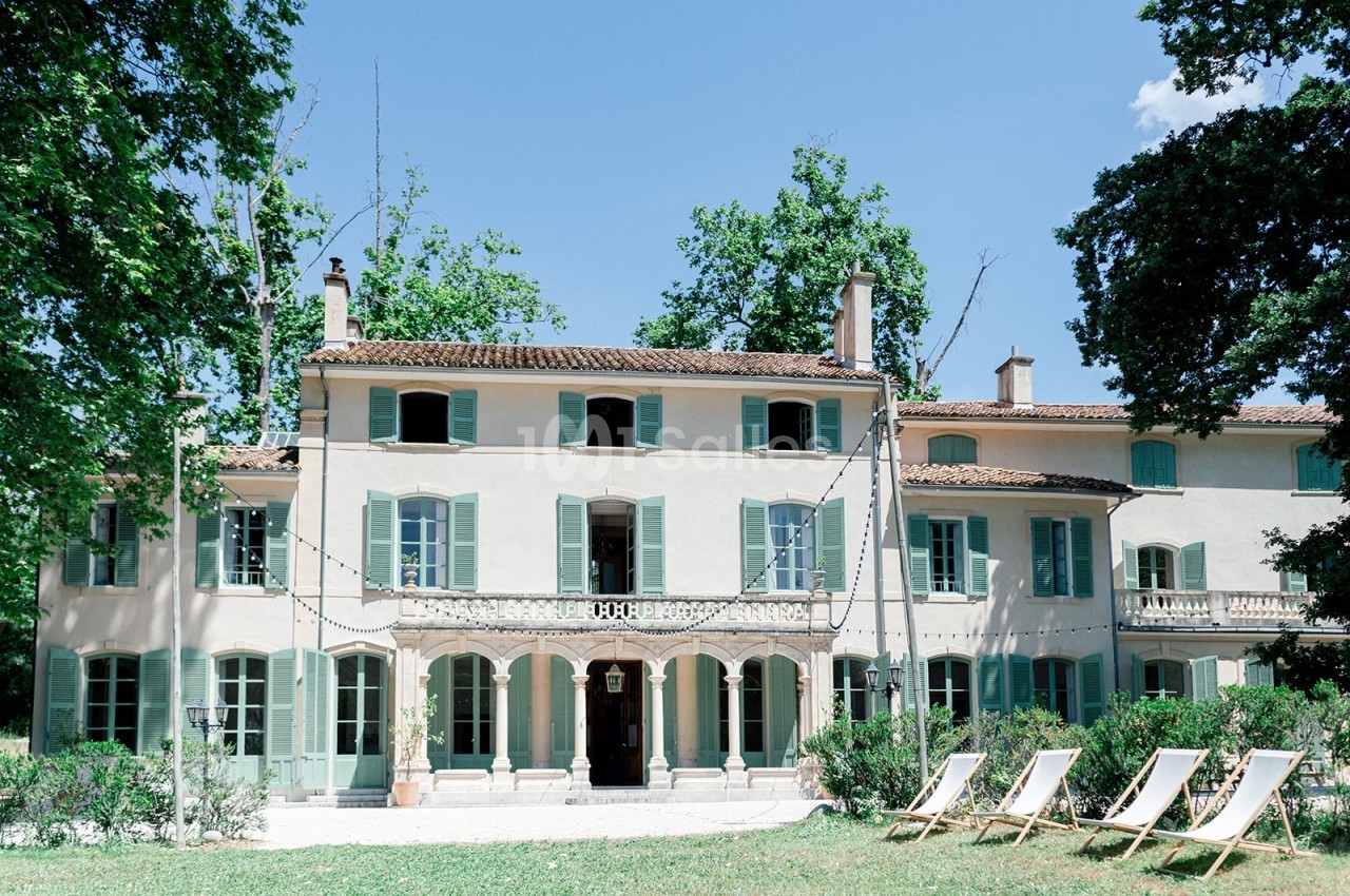 Façade d'une grande maison provençale avec volets verts, entourée de végétation et de chaises longues sur une pelouse.