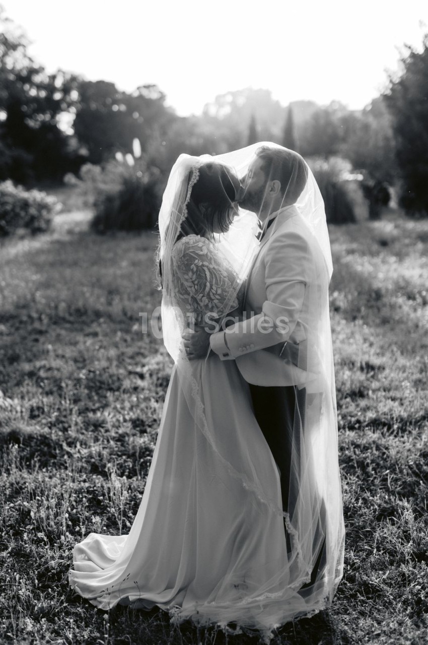 Un couple en tenue de mariage s'embrasse tendrement dans un champ, enveloppé d'un voile.