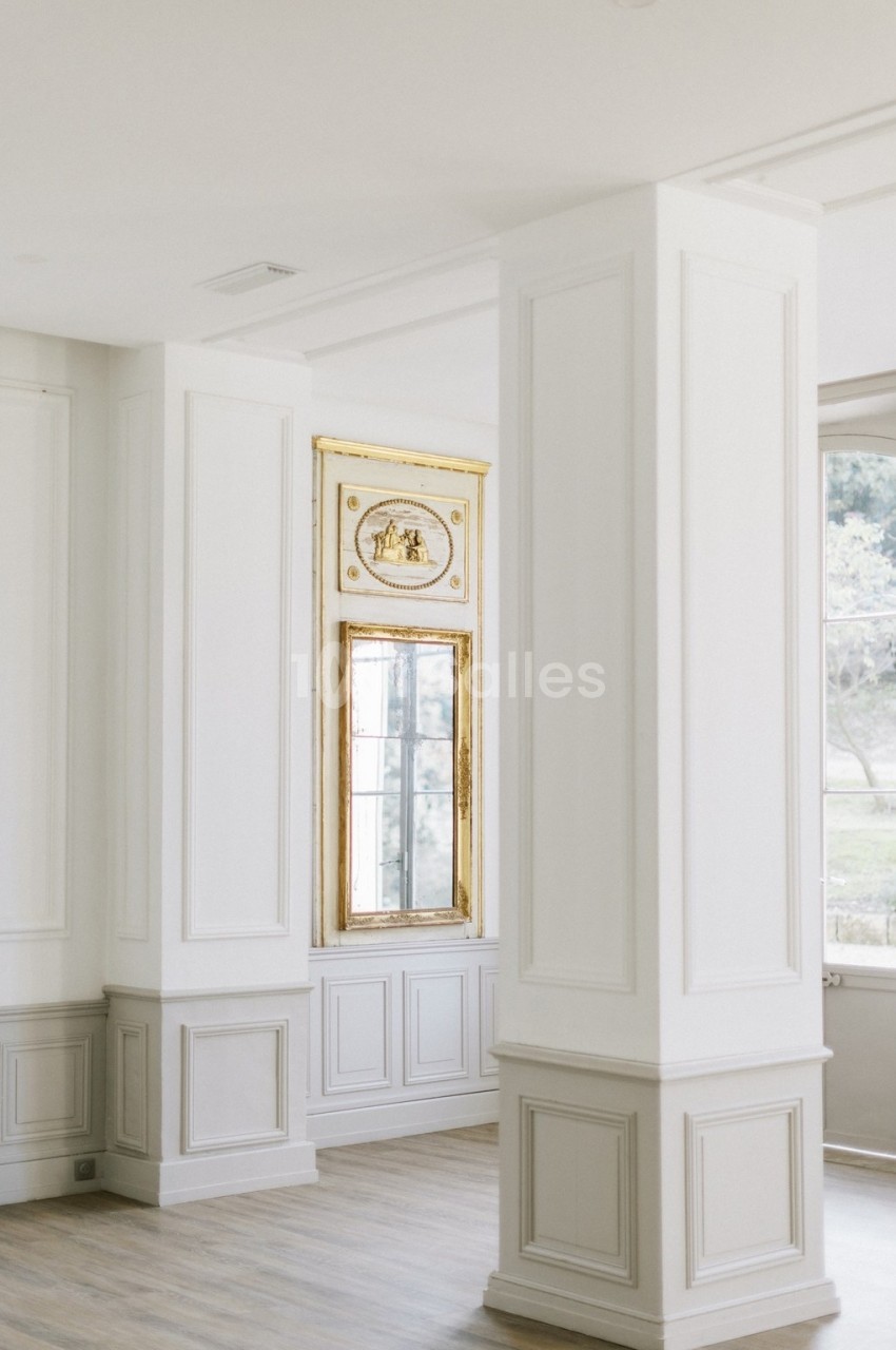Intérieur lumineux avec colonnes blanches, moulures décoratives et fenêtre encadrée de dorures donnant sur un espace…