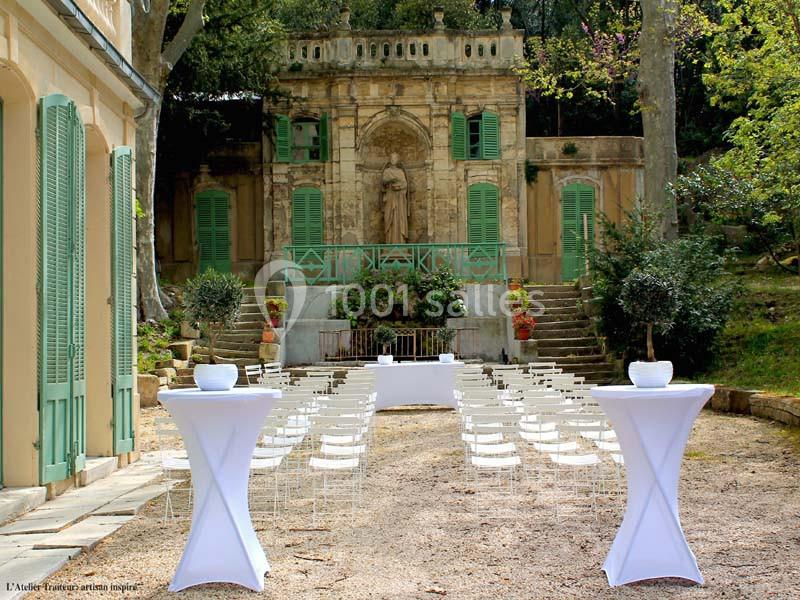 Chaises blanches alignées devant une table dans une cour extérieure, entourée de verdure et d'une façade ancienne.
