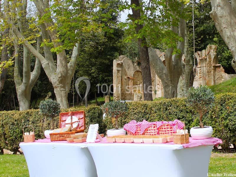 Table de buffet en plein air avec nappes blanches, paniers en osier et plantes, dans un parc arboré près de ruines anciennes.