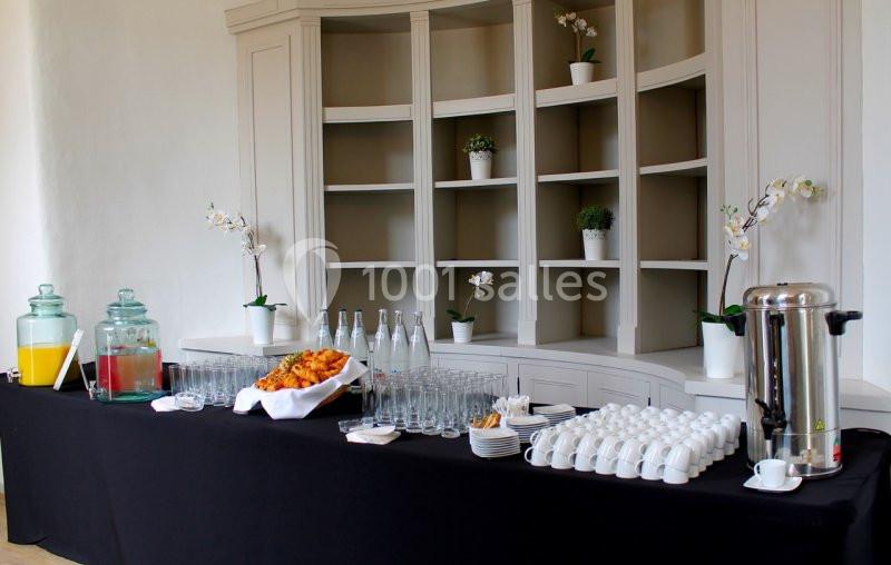 Table de buffet avec boissons, viennoiseries, tasses et verres devant une étagère décorée de plantes.