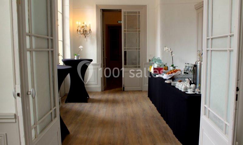 Salle lumineuse avec tables hautes et buffet comprenant boissons, viennoiseries et fleurs blanches en décoration.