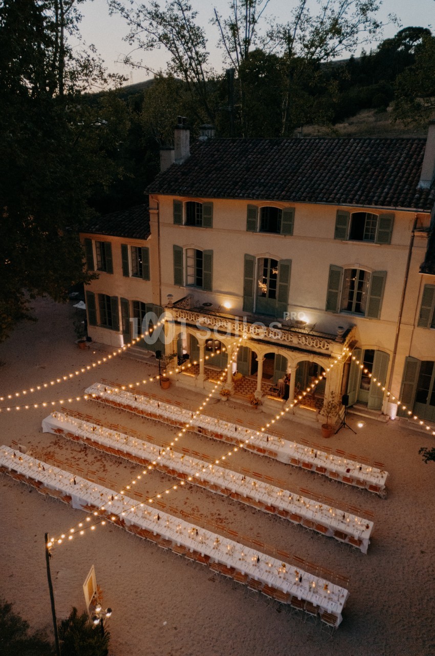 Grande maison avec tables longues décorées pour un dîner en extérieur, éclairées par des guirlandes lumineuses au crépuscule.