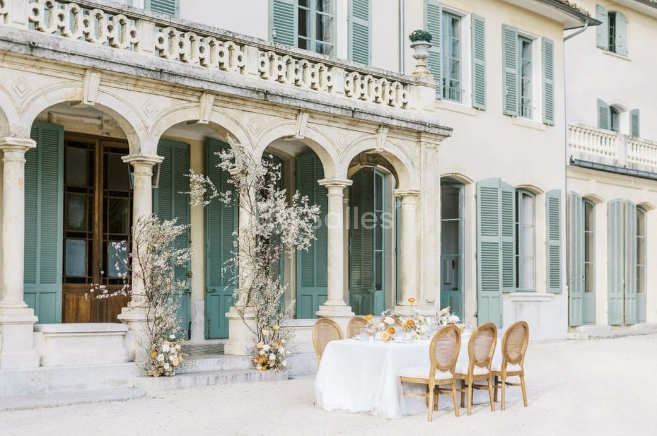 Table dressée en extérieur devant une maison aux volets verts, entourée d'arches et de décorations florales.