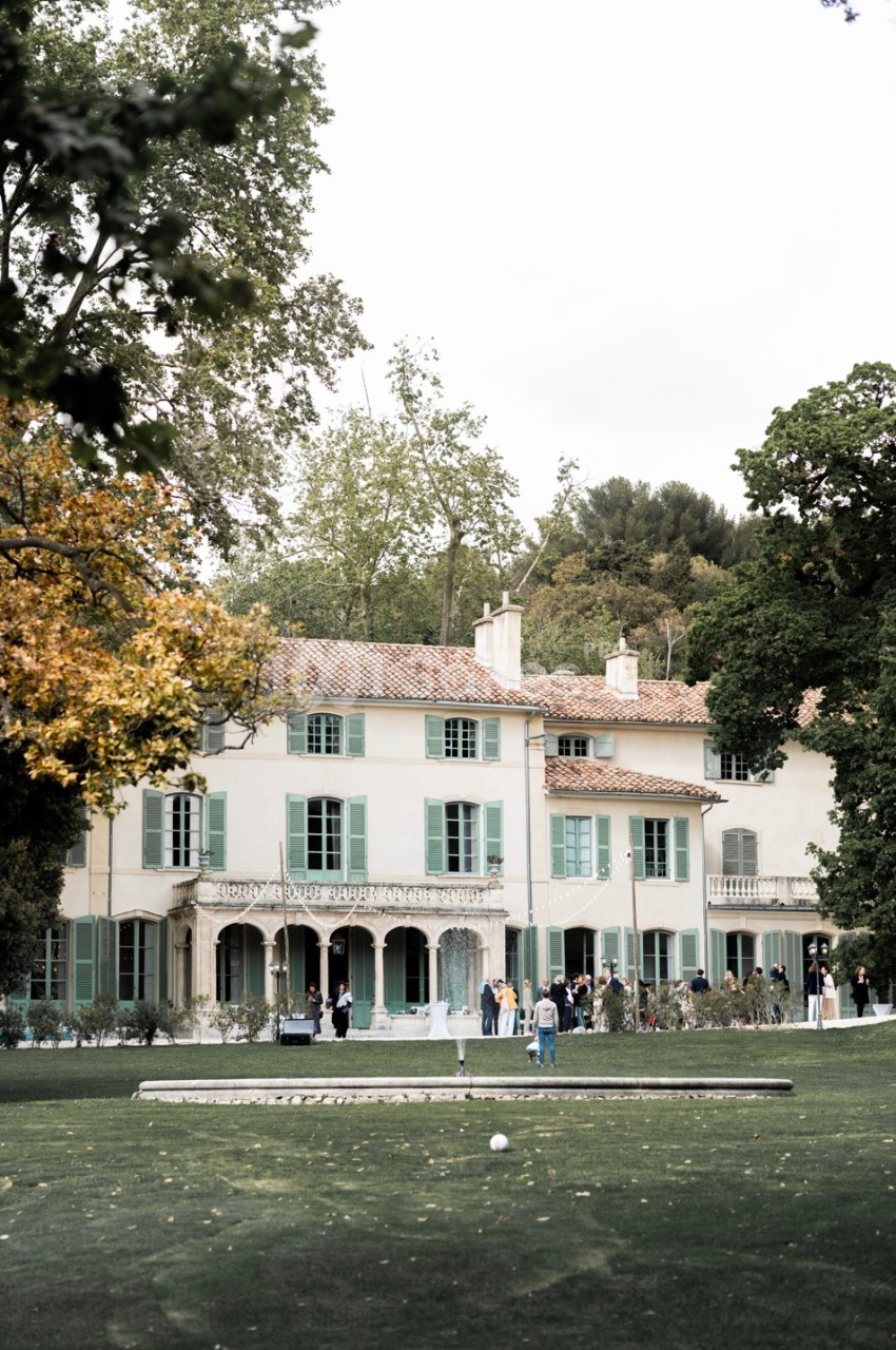 Grande maison blanche à volets verts entourée d'arbres, avec des personnes rassemblées sur la pelouse devant.