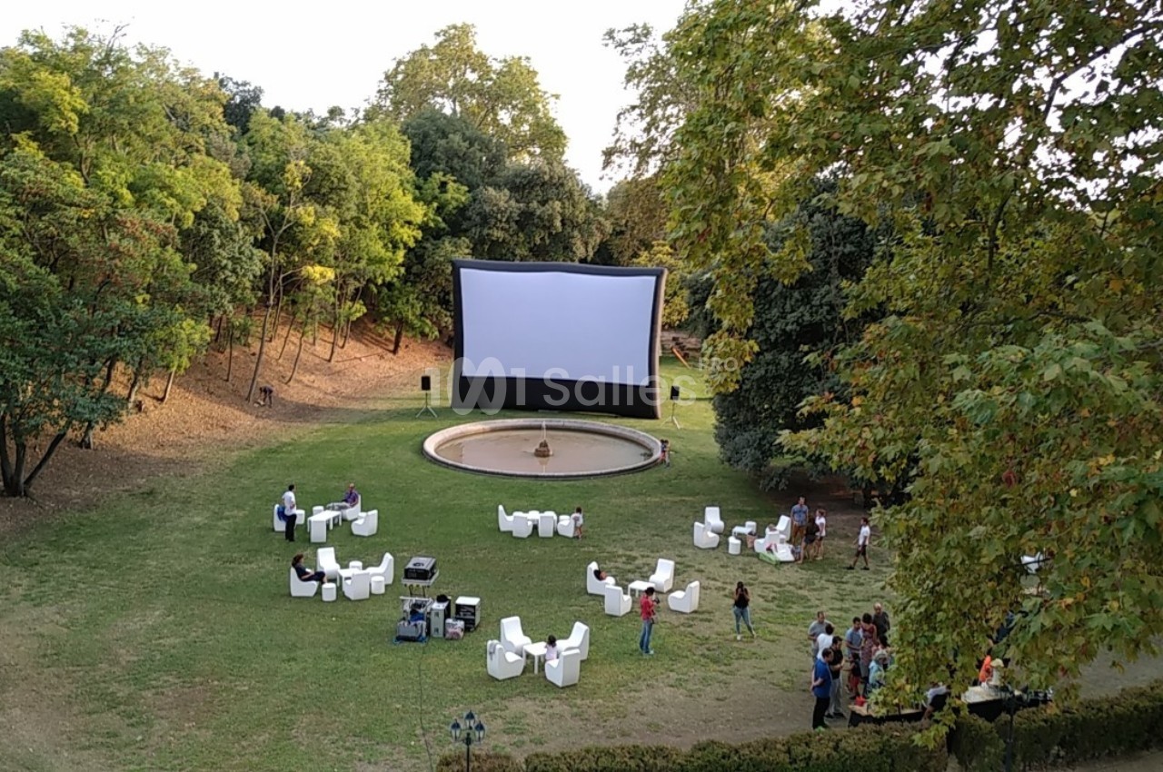 Écran de projection en plein air installé dans un parc, entouré de chaises blanches et de quelques personnes.