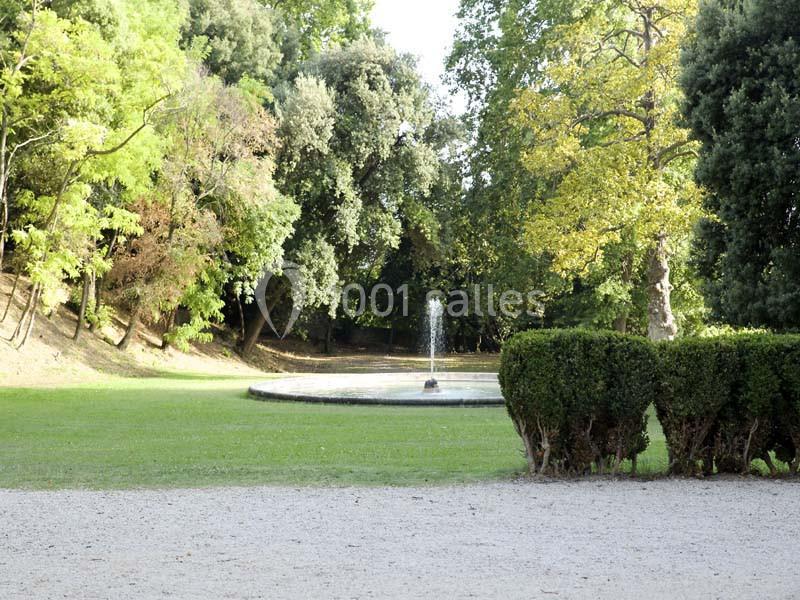 Fontaine entourée de pelouse et d'arbres dans un parc, avec une haie basse au premier plan.