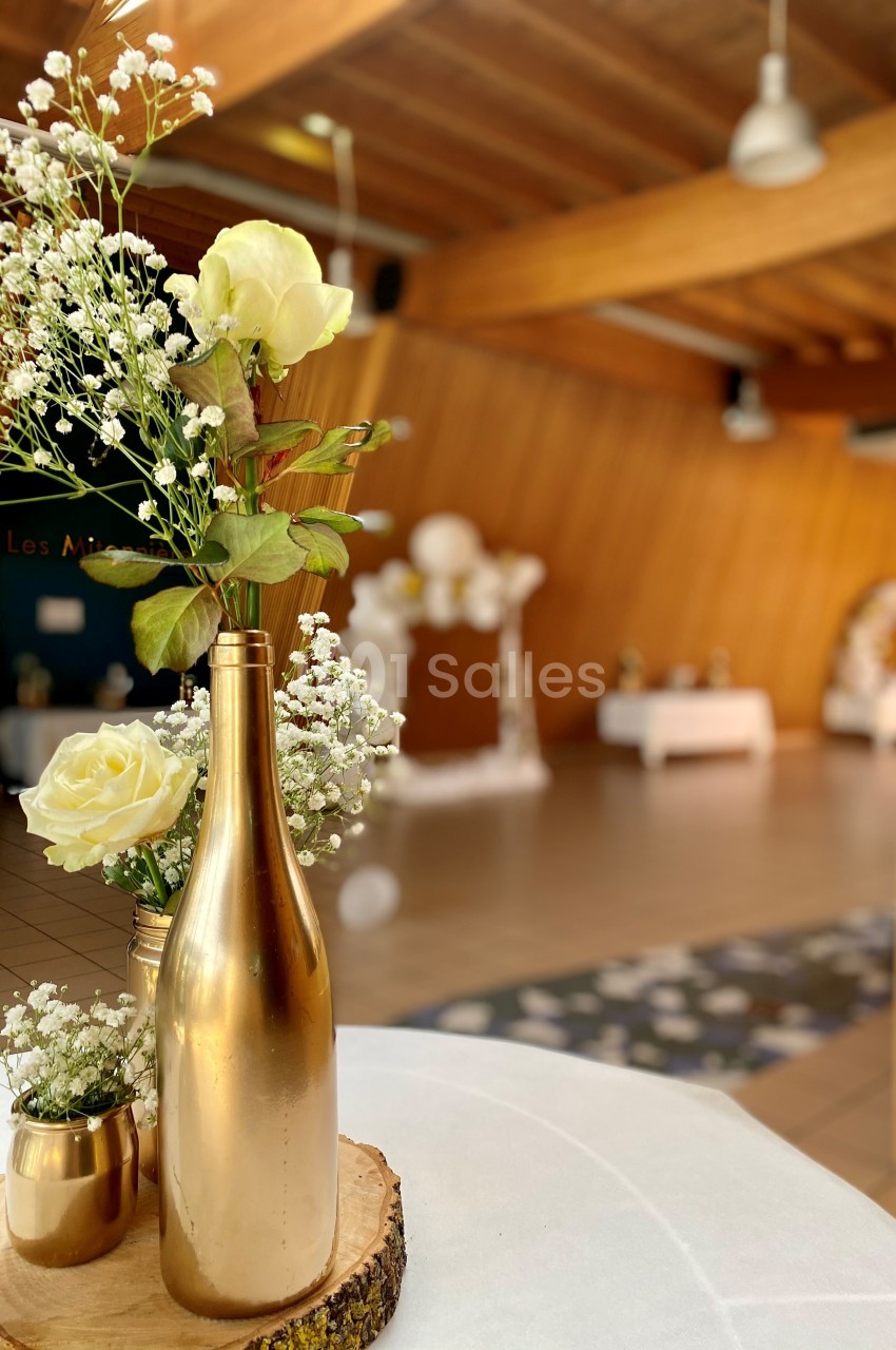 Centre de table avec bouteille dorée, fleurs blanches et jaunes, dans une salle en bois décorée pour un événement.