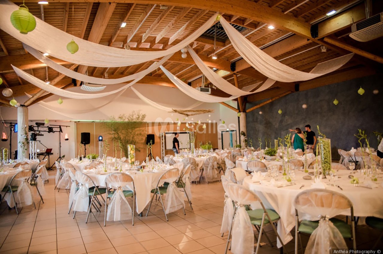 Salle de réception décorée avec des tables rondes, nappes blanches, chaises habillées et voilages suspendus au plafond.