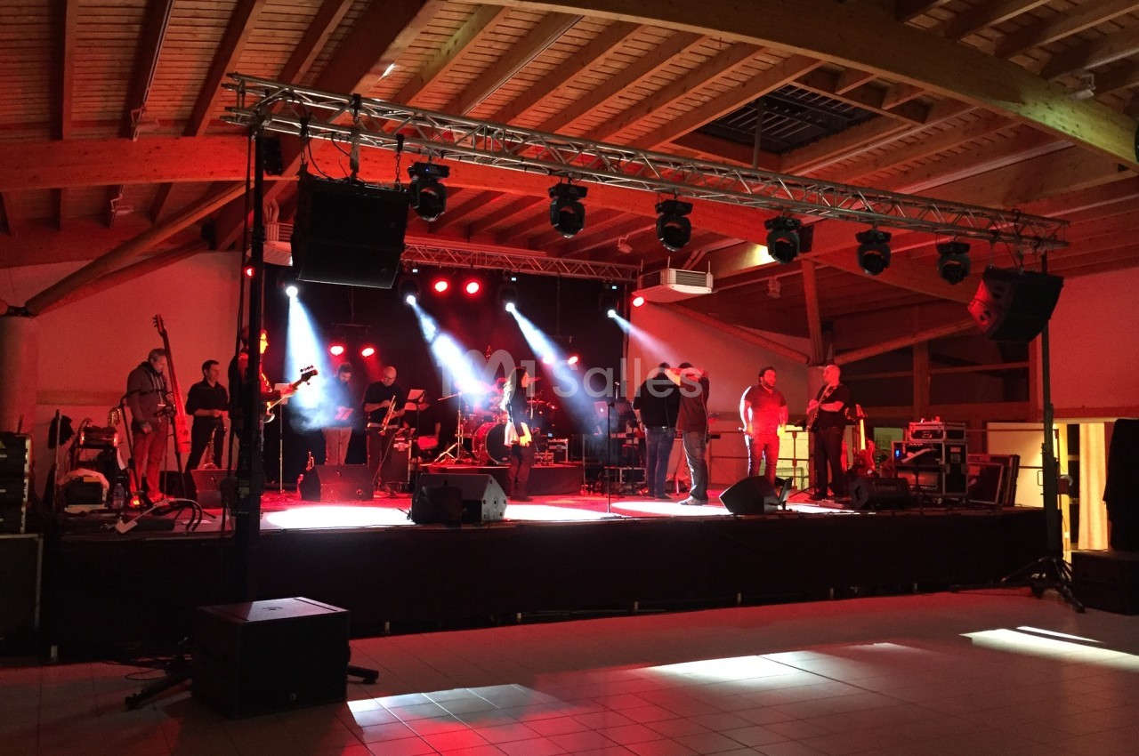 Groupe de musiciens sur une scène éclairée par des projecteurs rouges dans une salle avec charpente en bois.
