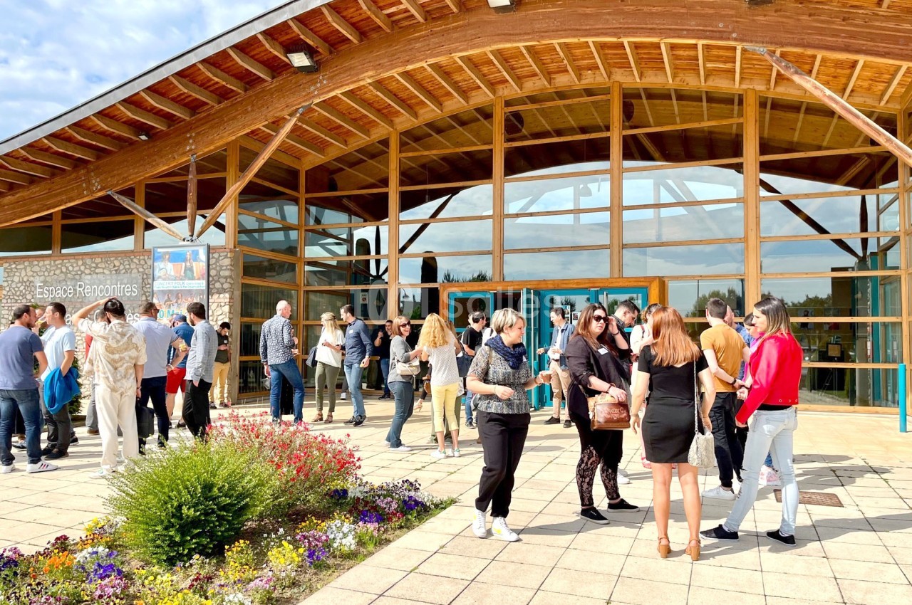 Groupe de personnes discutant devant un bâtiment en bois avec une façade vitrée, entouré de fleurs et d'une terrasse pavée.